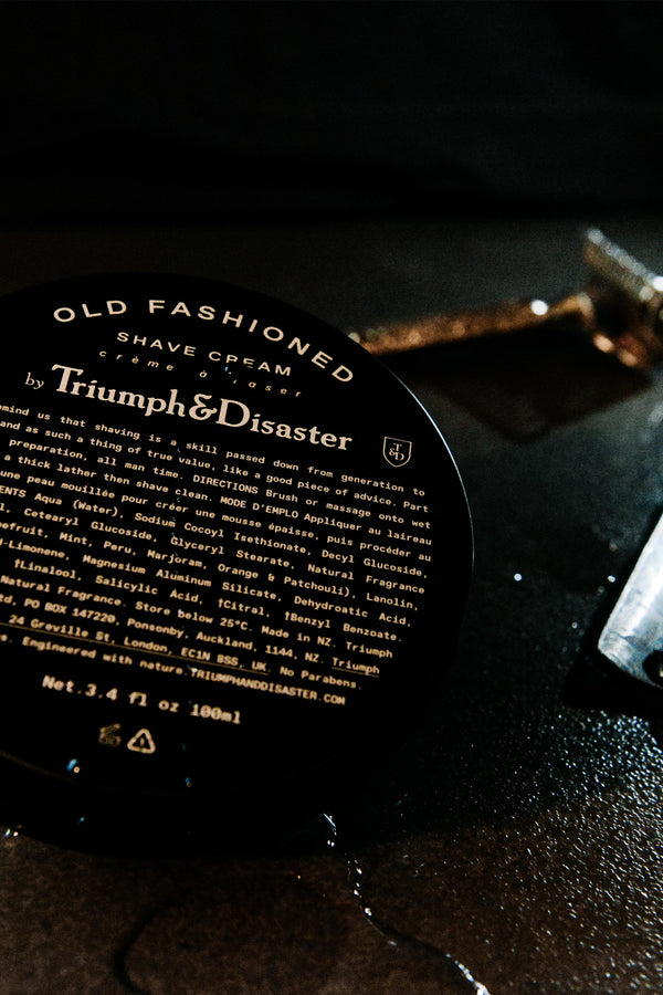 A close-up of the Triumph & Disaster Old Fashioned Shave Cream Jar rests on a dark surface, with a gold razor and a glass object softly blurred in the background, capturing traditional shaving style.