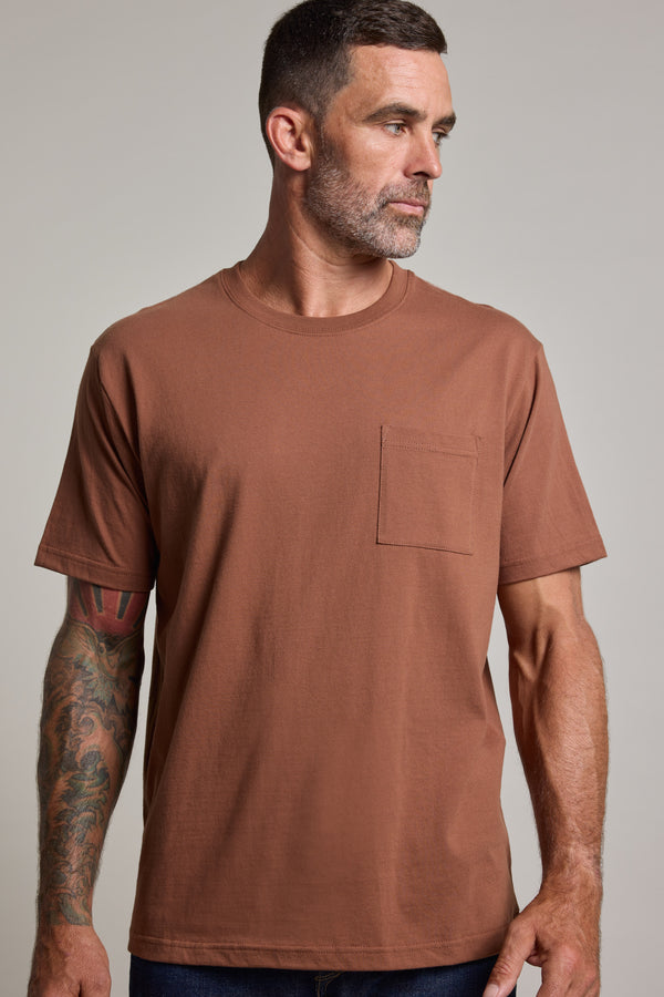 A man with short dark hair and a beard, wearing the Barkers Wrenches Print Tee—a brown heavyweight t-shirt with a chest pocket—stands against a plain background, looking left. His right arm features colorful tattoos.
