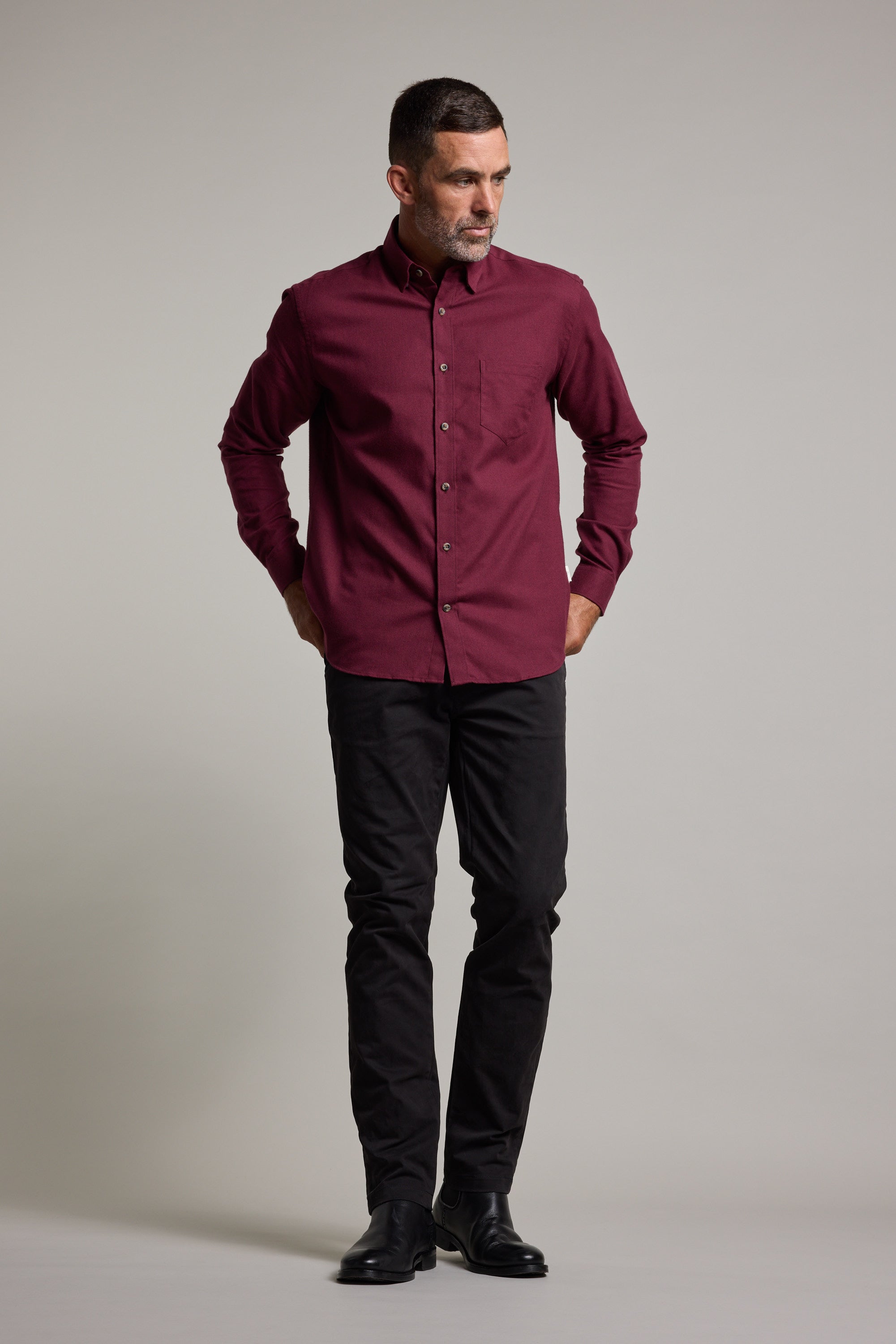 A man stands against a plain background in the Barkers Antonio Shirt in maroon, made from a soft cotton-wool blend. He pairs it with black pants and shoes, gazing to his right with his hands in his pockets.