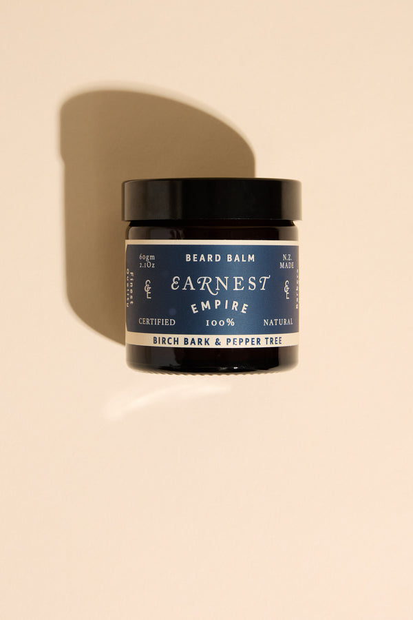 A black jar of Other Earnest Empire Beard Balm with a blue label sits on a beige surface, casting a shadow. The label features Birch Bark and Pepper Tree scent, natural ingredients, and an NZ Made certification.