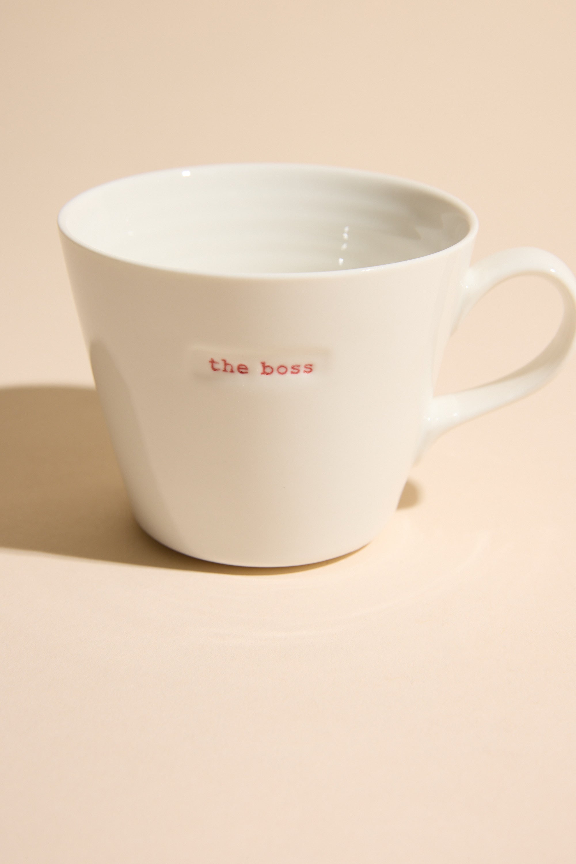 The "Bucket Mug - The Boss" by Other is a white ceramic mug with "the boss" in red, displayed on a light beige surface under soft lighting—a stylish designer homeware piece.
