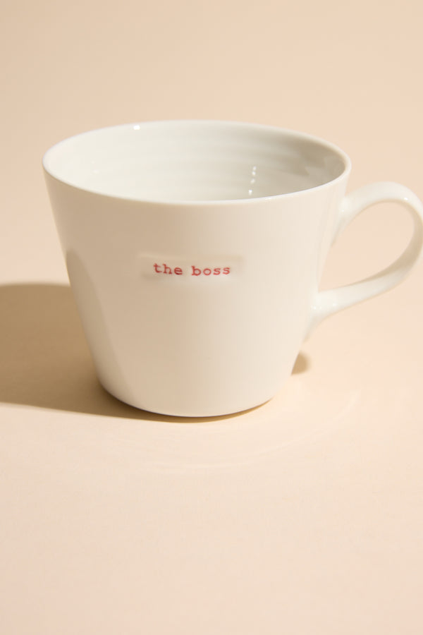 The "Bucket Mug - The Boss" by Other is a white ceramic mug with "the boss" in red, displayed on a light beige surface under soft lighting—a stylish designer homeware piece.