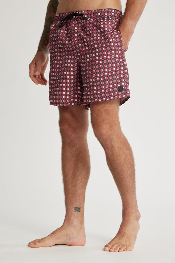 A person stands barefoot in Barkers’ Laramie Print Volley maroon shorts with a pink geometric pattern. Crafted from recycled polyester with a drawstring waist, the quick-drying shorts reveal tattoos, shown against a plain white background.