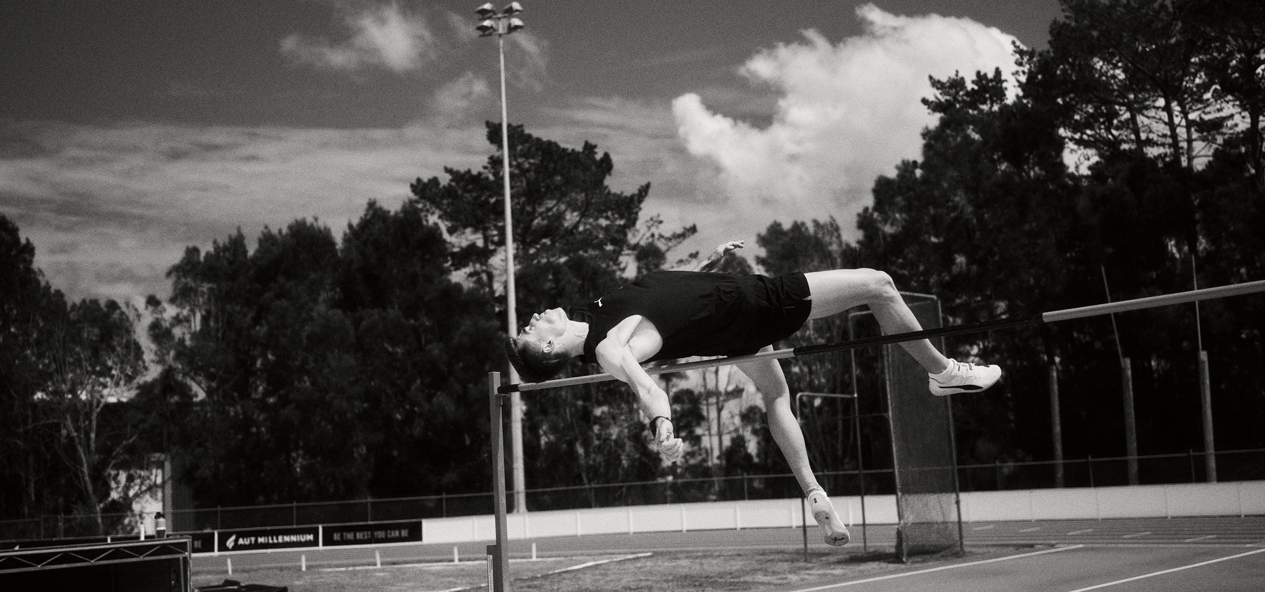 NZ athlete in mid-air clearing a high jump bar on an outdoor track.