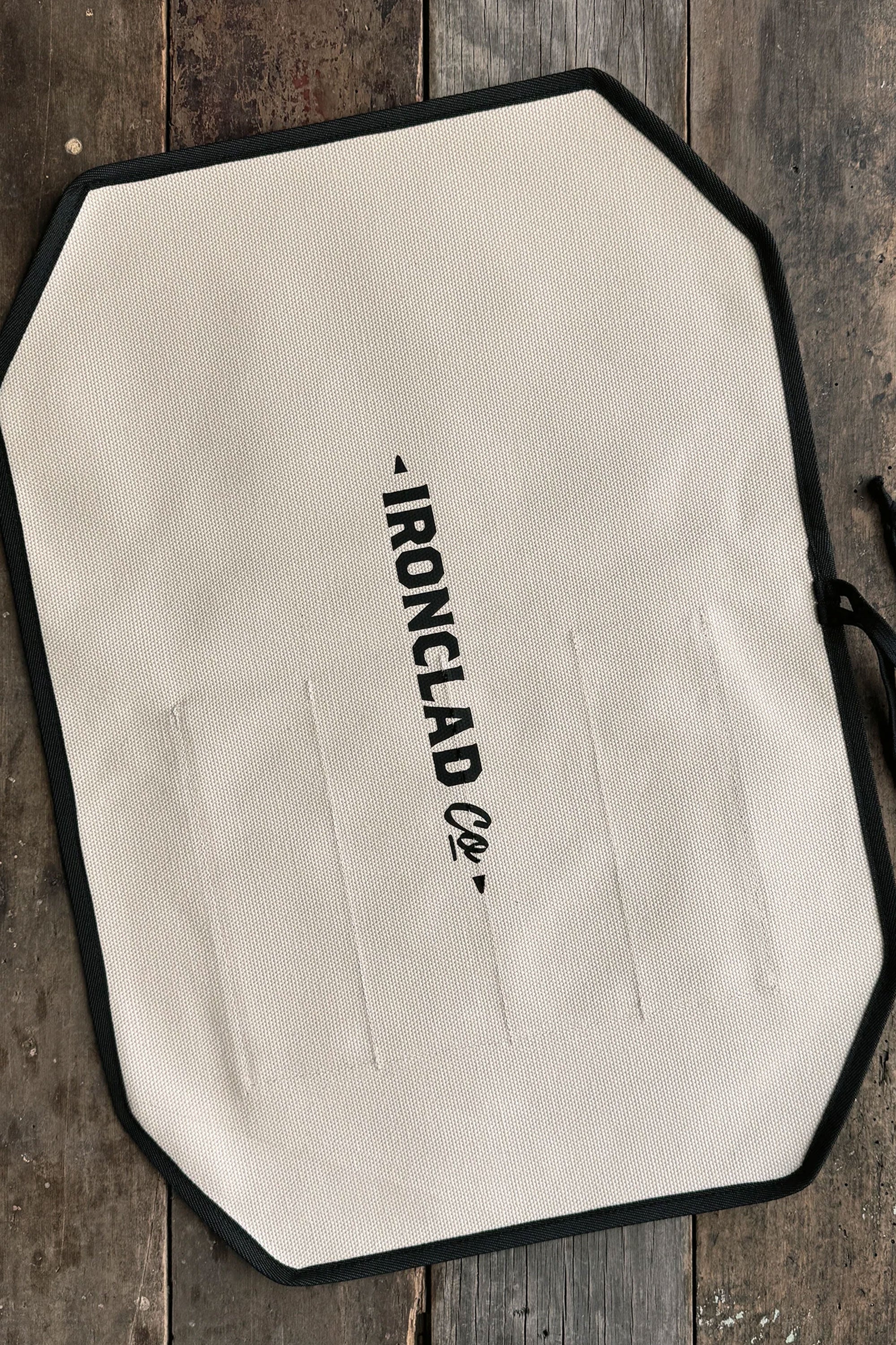 A white, octagonal IronClad apron with black trim and logo lies flat on a rustic wooden surface beside an IronClad Ghost Knife Roll.