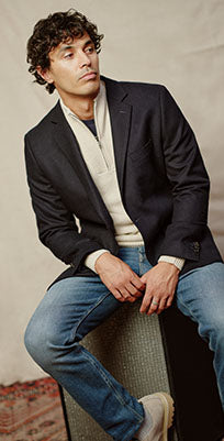 A man with curly hair wearing a navy blazer, cream sweater, blue jeans, and beige shoes sits on a patterned box, looking to the side against a neutral background.