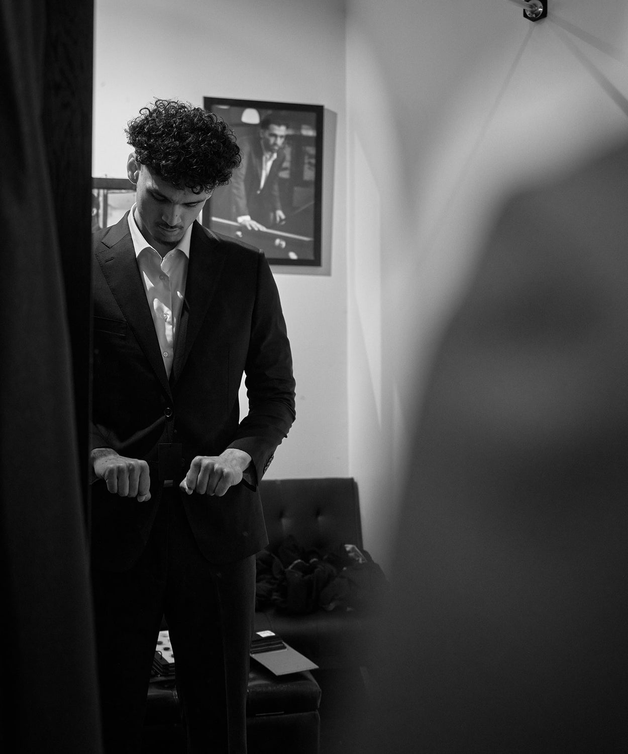 Black and white photo of a young man with curly hair adjusting his suit jacket in a room, with a framed picture of another man in a suit on the wall behind him.