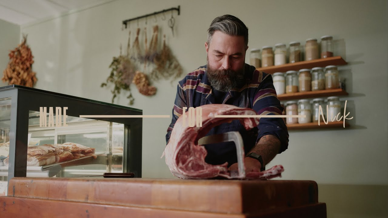 A bearded man slices NZ meat in a butcher shop with jars on shelves behind him.
