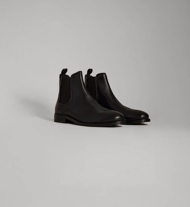 A pair of black leather Chelsea boots on a plain light gray NZ background.
